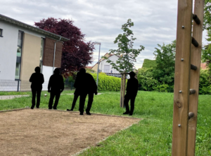 Silhouetten von vier Personen beim Boule-Spiel auf einem sandigen Spielfeld im Freien, umgeben von grünem Rasen, Bäumen und einem Gebäude im Hintergrund.